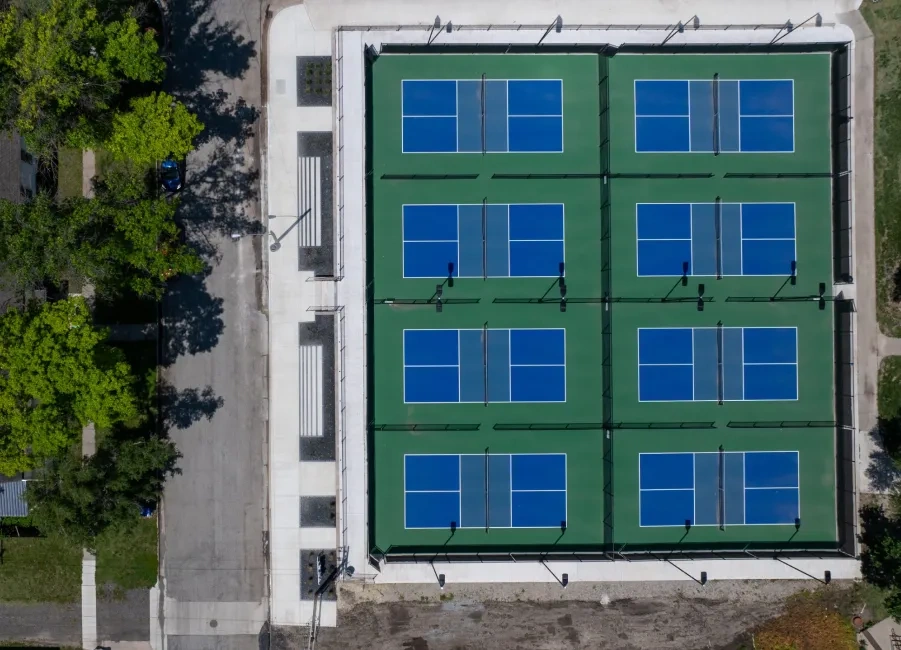 A straight-down view of eight blue and green pickleball courts at McPherson Commons, enclosed by black fencing, with a poured concrete apron and steps along one side.