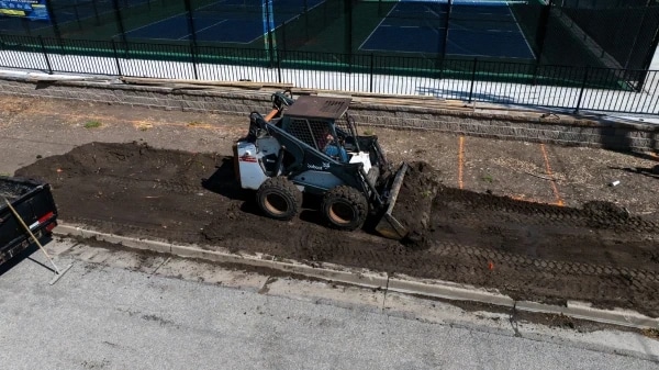A skid steer grading the parkway strip from above, the block wall and courts fence behind it and fresh soil ridged where the beds will go.