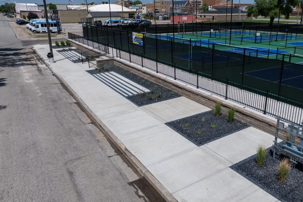 The street edge of the McPherson courts: new concrete walk and steps, black-rock beds with young grasses set into the paving, and the fenced courts above.