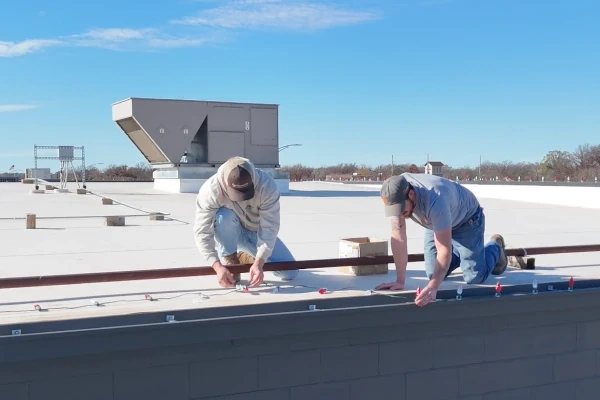Two of the crew kneeling on the white commercial roof in daylight, fixing the light track along the parapet edge.