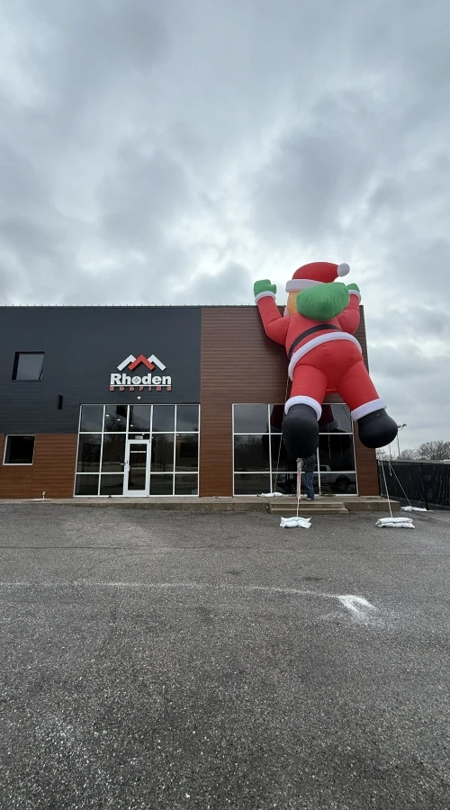 An inflatable Santa mounted on the roof of the Rhoden Roofing building above the car park, under an overcast sky.