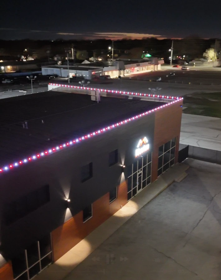 The Rhoden Roofing building after dark with the roofline picked out in red and white lights, the lit signage below it and the town behind.