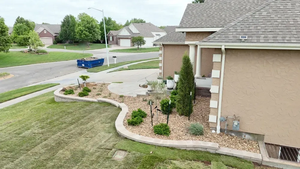 The raised planter at the Riley residence: a curved block wall holding decorative rock, boulders and young shrubs, with the bed running on along the side of the house.