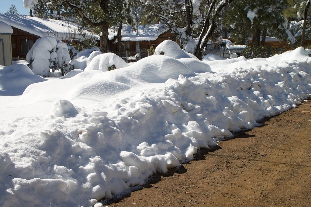A long bank of plowed snow piled along a cleared lane, with snow-covered houses and trees behind it.