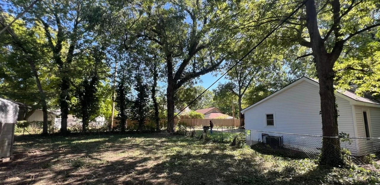 The same lot cleared to open ground under the mature trees, a white garage and a chain-link fence in view at the side.