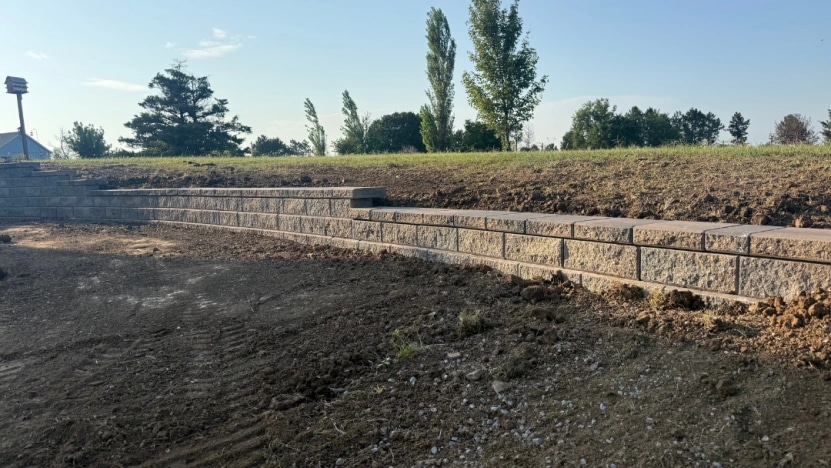 A two-tier segmental block retaining wall stepping along a freshly graded slope, bare soil in the foreground and new grass rising behind it.