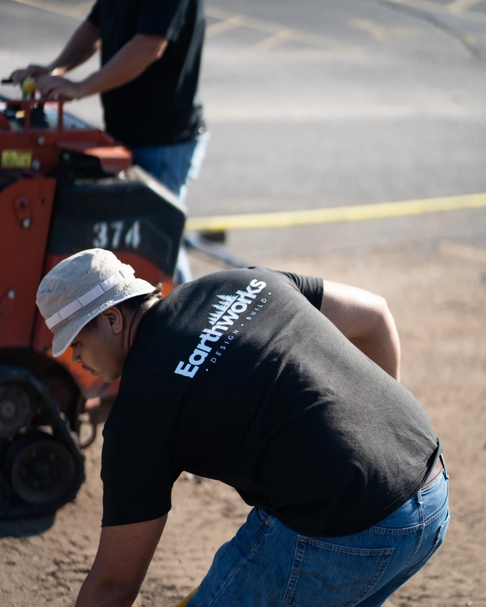 A crew member in an Earthworks shirt and bucket hat bent to the ground beside a walk-behind trencher, a second at its controls behind him.